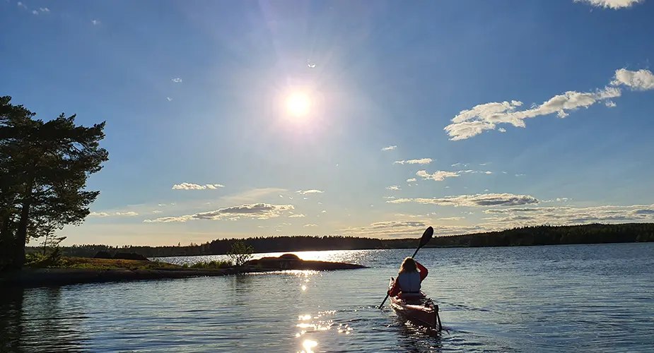 Kvinna paddlar kajak på spegelklart vatten. Stor sol och blå himmel