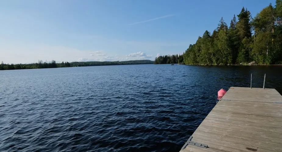 Blå sjö med skog i bakgrunden och en träbrygga i högerkant