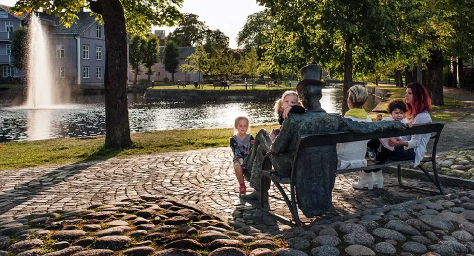 Ferlinstatyn i Filipstads centrum tillsammans med tre småbarn och två vuxna som sitter på parkbänken med fontänen i bakgrunden