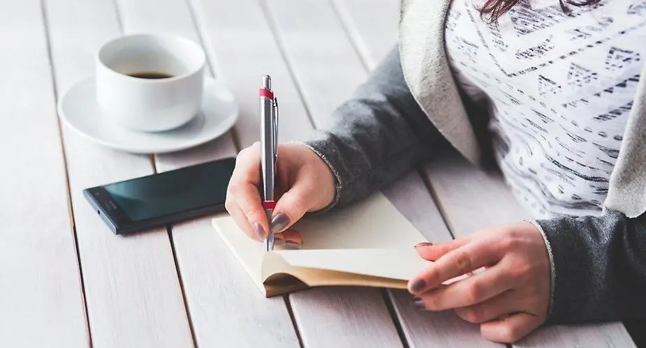 Kvinna med mobiltelefon och kaffekopp skriver anteckningar vid vitt träbord 
