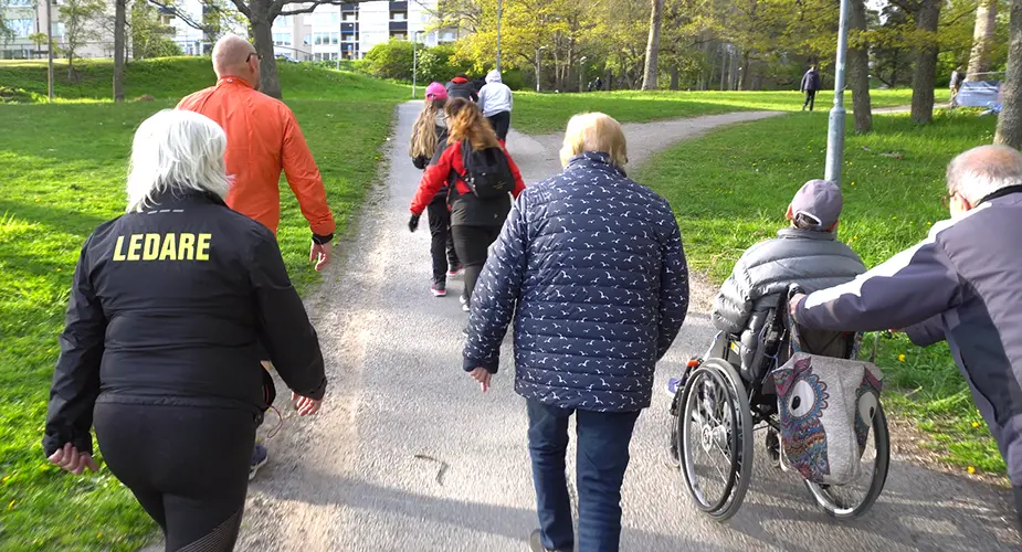 En grupp personer promenerar uppför en asfalterad väg, en rullar en person i rullstol