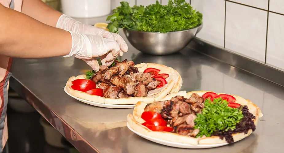 Kebab upplagt fint på två tallrikar på en bänk i ett kök, kocken har plasthandskar