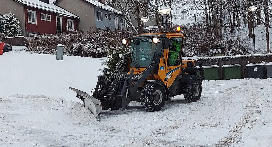 Gul traktor plogar snö med tre hus i bakgrunden
