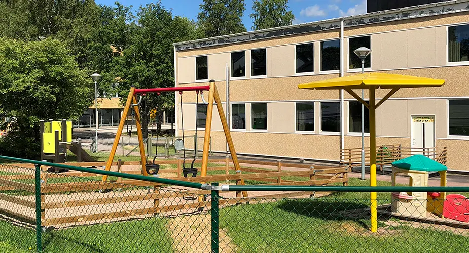 Förskolan Lyckebo. Beige stenhus, två våningar, med en lekpark utanför med gungor och liten klätterställning, gröt staket runt hela lekparken.